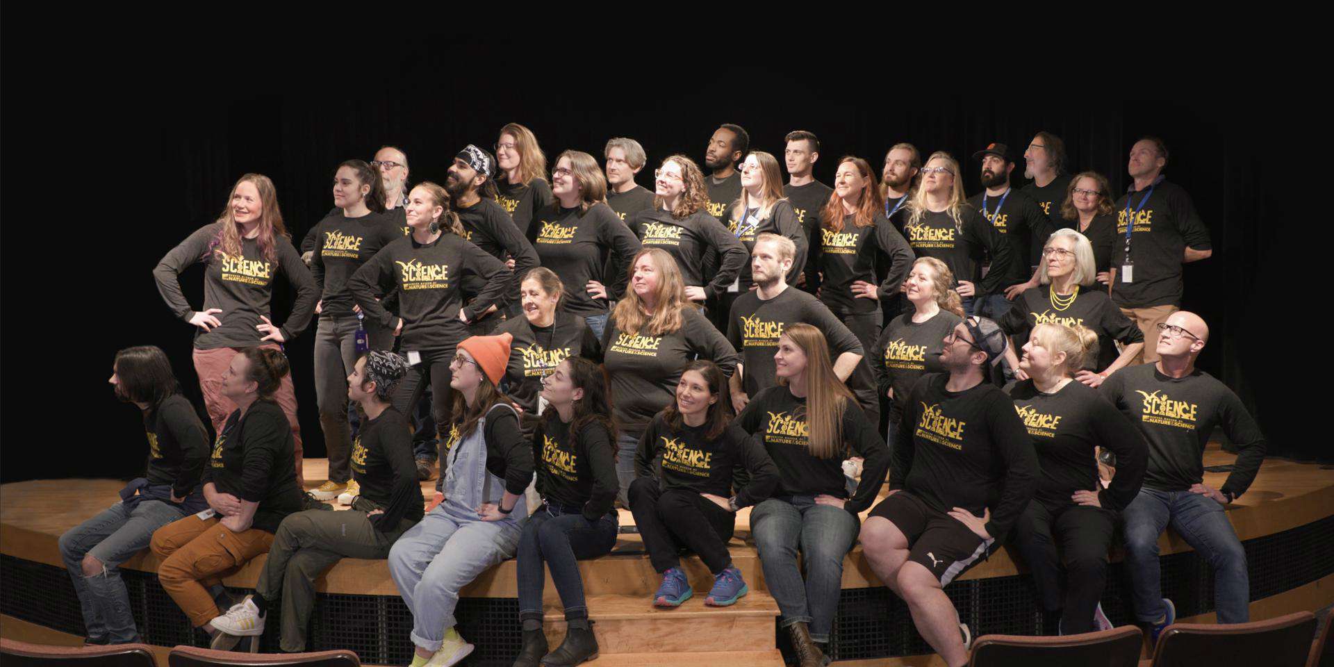 a large group of staff all wearing Science Division shirts stand on a stage with their hands on their hips looking off to the right.