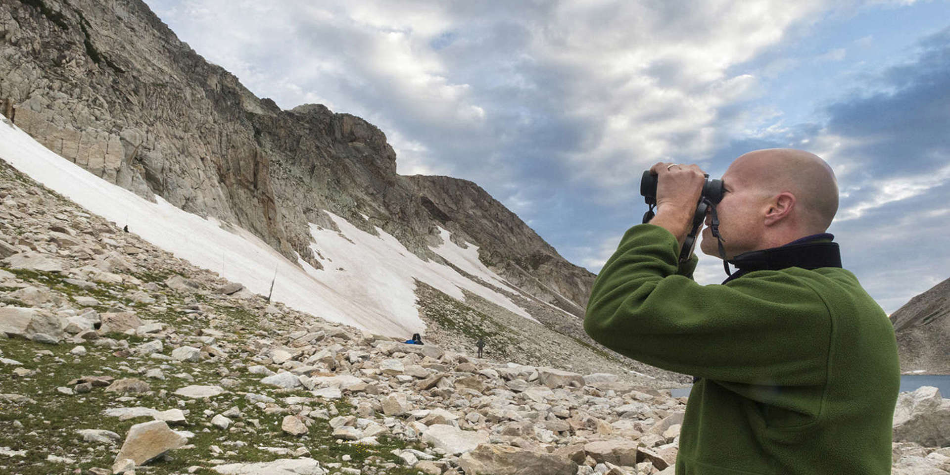 Dr. Garth Spellman stands looking through binoculars at a large snow-covered cliff